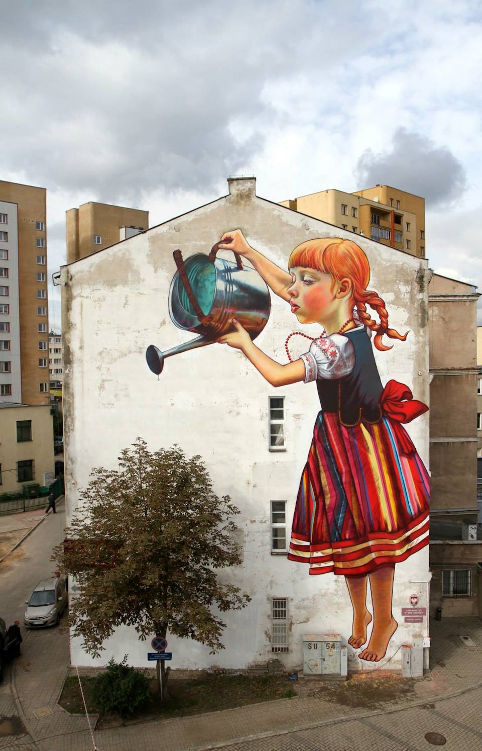 Mural of a girl in traditional clothing realistically painted watering an actual tree positioned next to the mural on a building wall.