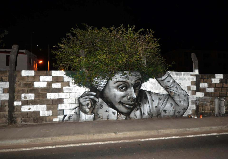Mural painting of a figure combing their hair, using real bushes as their hair on a street wall.