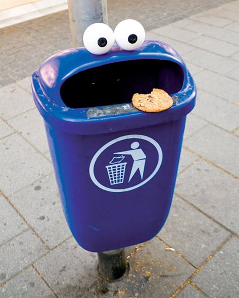 Purple public trash bin on a sidewalk with two large googly eyes on top and a cookie on the rim, making the bin look like Cookie Monster.