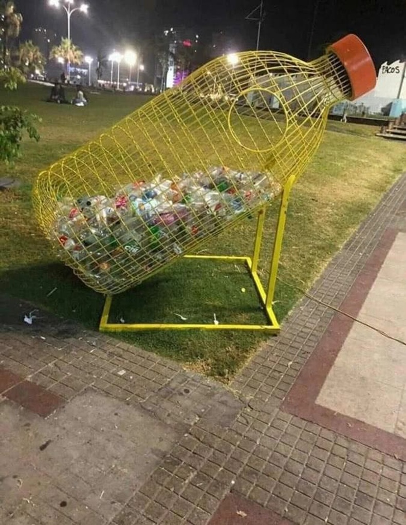 Yellow wire-frame bottle-shaped recycling bin filled with plastic bottles on grass beside a sidewalk at night.