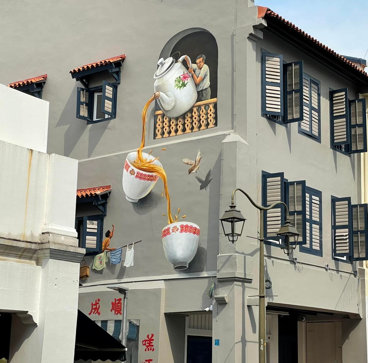 A hyperrealistic mural by Singaporean artist Yip Yew Chong on the wall of a building at 30 Temple Street in Chinatown, Singapore. The artwork creates the illusion of a man pouring tea from a giant porcelain teapot into two oversized teacups suspended mid-air, with the tea stream forming a dynamic connection between them. A bird flies through the scene and a figure hangs laundry beneath the floating cups, integrating local daily life into the surreal composition. Painted in a trompe-l'œil style, the mural cleverly blends fantasy with heritage and celebrates traditional Kopitiam culture.