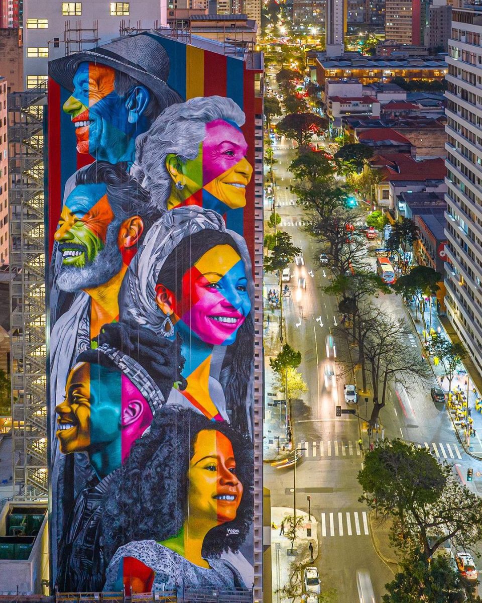 A massive Eduardo Kobra mural in Belo Horizonte, Brazil, showing colorful layered portraits on the side of a tall city building at night.