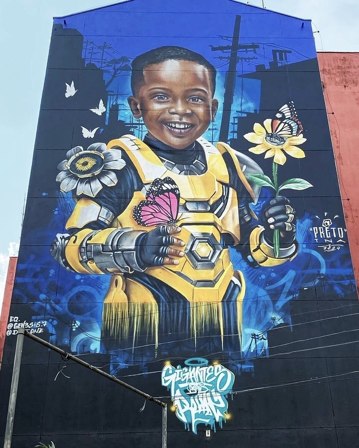 A tall mural by PRETO in Perus, São Paulo, Brazil, showing a smiling child in futuristic yellow armor holding flowers and butterflies against a blue background.