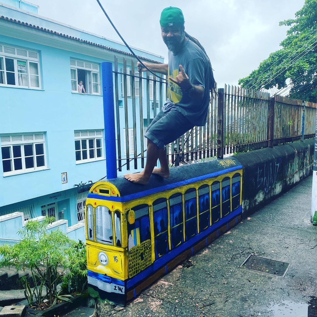 A wall intervention by André Kajaman in Rio de Janeiro, Brazil, showing a painted yellow Santa Teresa tram along a sloped wall with a person standing on it.
