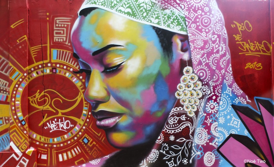 A mural by NOE TWO in Favela Vidigal, Rio de Janeiro, Brazil, showing a colorful side-profile portrait with ornate patterns and a red background.