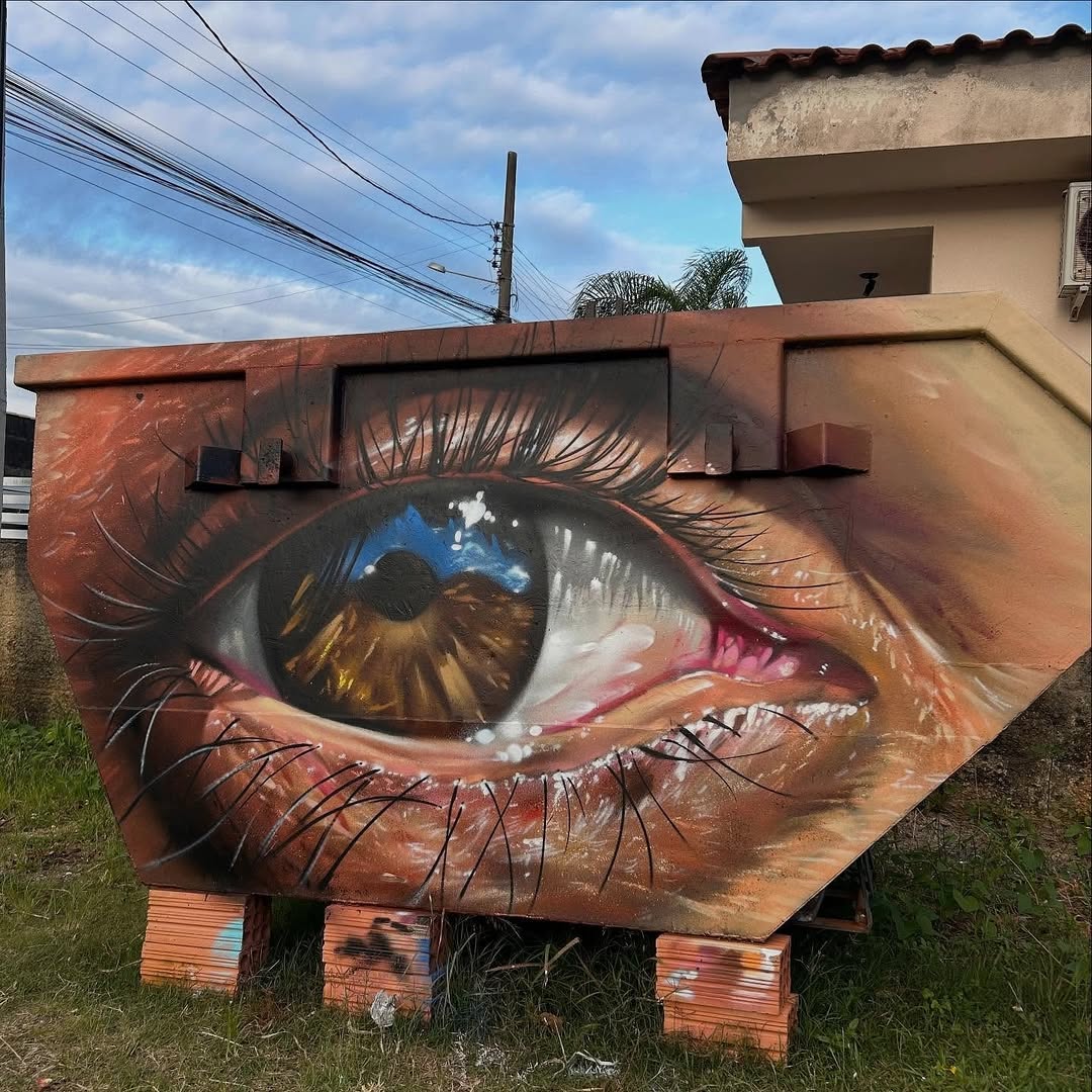 Artwork by HEROK in Balneário Rincão, Brazil showing a hyper-realistic human eye with a brown iris, painted across the surface of a large metal dumpster, complete with detailed lashes and skin texture.