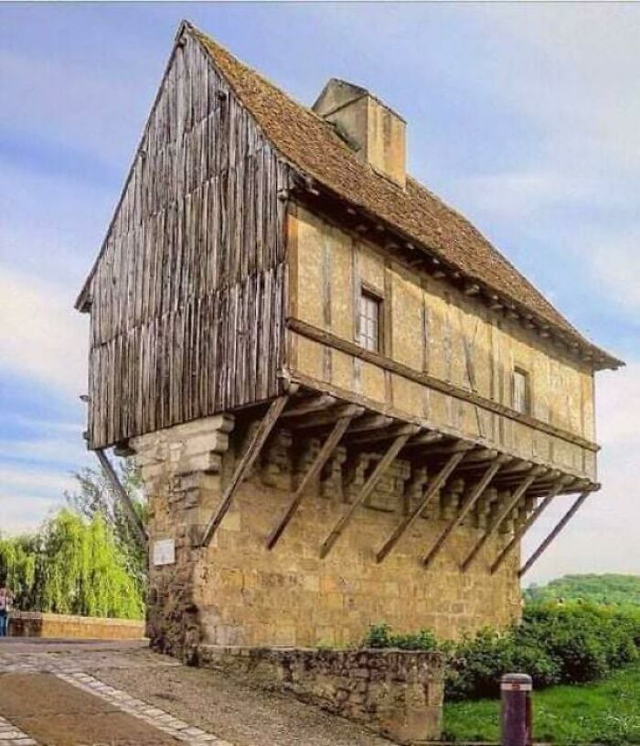 Historic timber house perched on stone supports with angled wooden braces, creating an overhang that looks precariously balanced.
