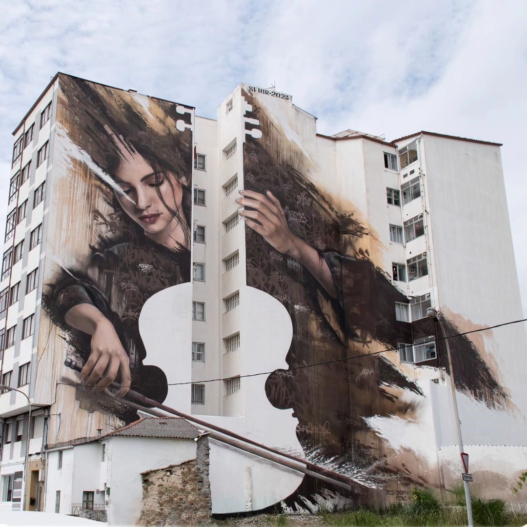 A monumental mural by SFHIR in Fene, Spain, depicting a woman playing a cello. The artwork spans multiple building faces, creating a stunning visual of musical grace with rich textures and lifelike details. It showcases SFHIR's mastery of scale and realism in street art.