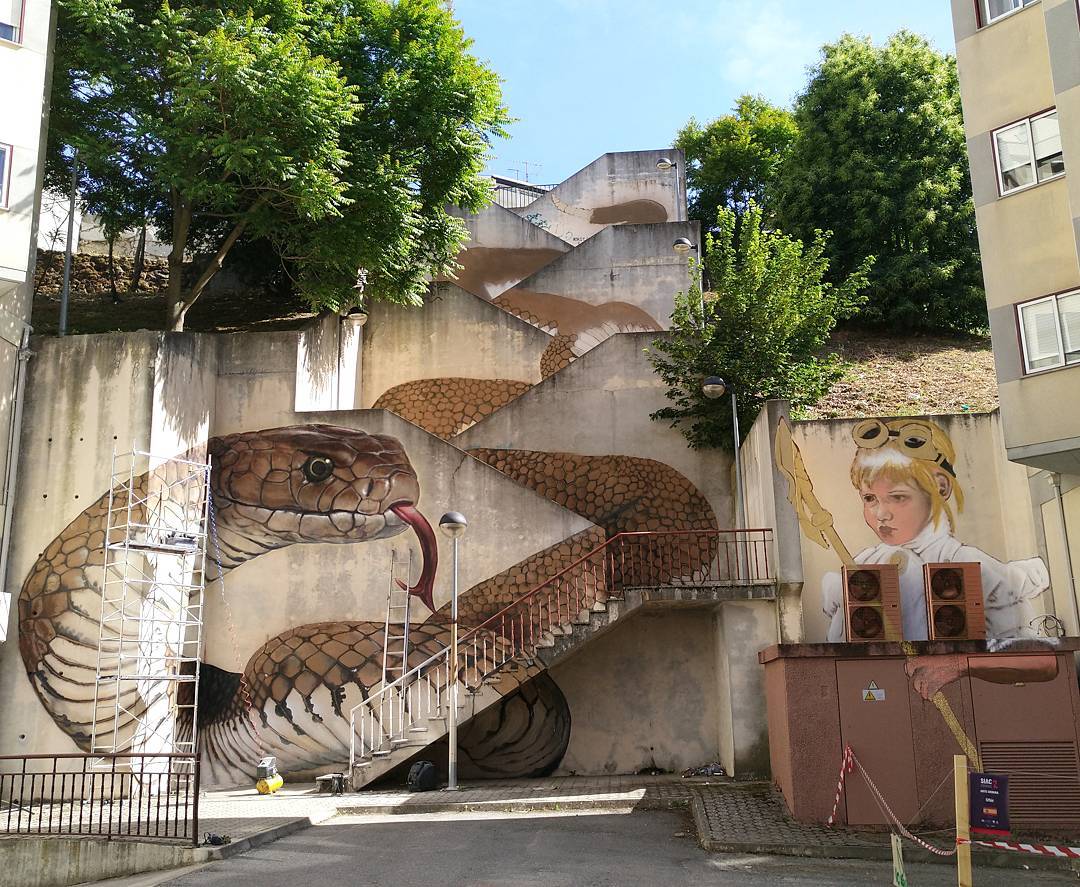 "Snake Stairs" by SFHIR in Guarda, Portugal. The mural transforms a staircase into a coiled snake, using the steps and walls to create the illusion of a slithering serpent. Beside the snake, a child dressed as an adventurer holds a staff, adding a sense of storytelling and exploration to the urban space.