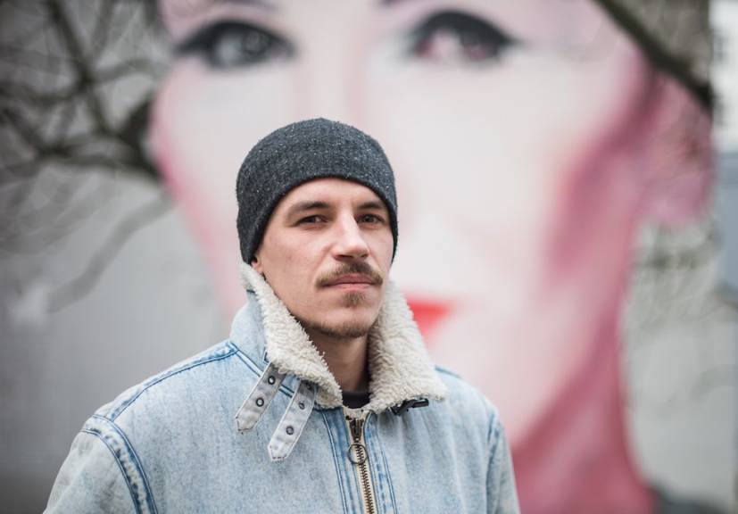 A portrait of street artist Bruno Althamer standing in front of his mural of Polish singer Kora (Olga Jackowska) in Warsaw, Poland. Althamer is wearing a black beanie and a denim jacket with a shearling collar, looking confidently at the camera. The mural, featuring Kora's face with red lips and expressive eyes, serves as the background, blending seamlessly with the tree's branches.