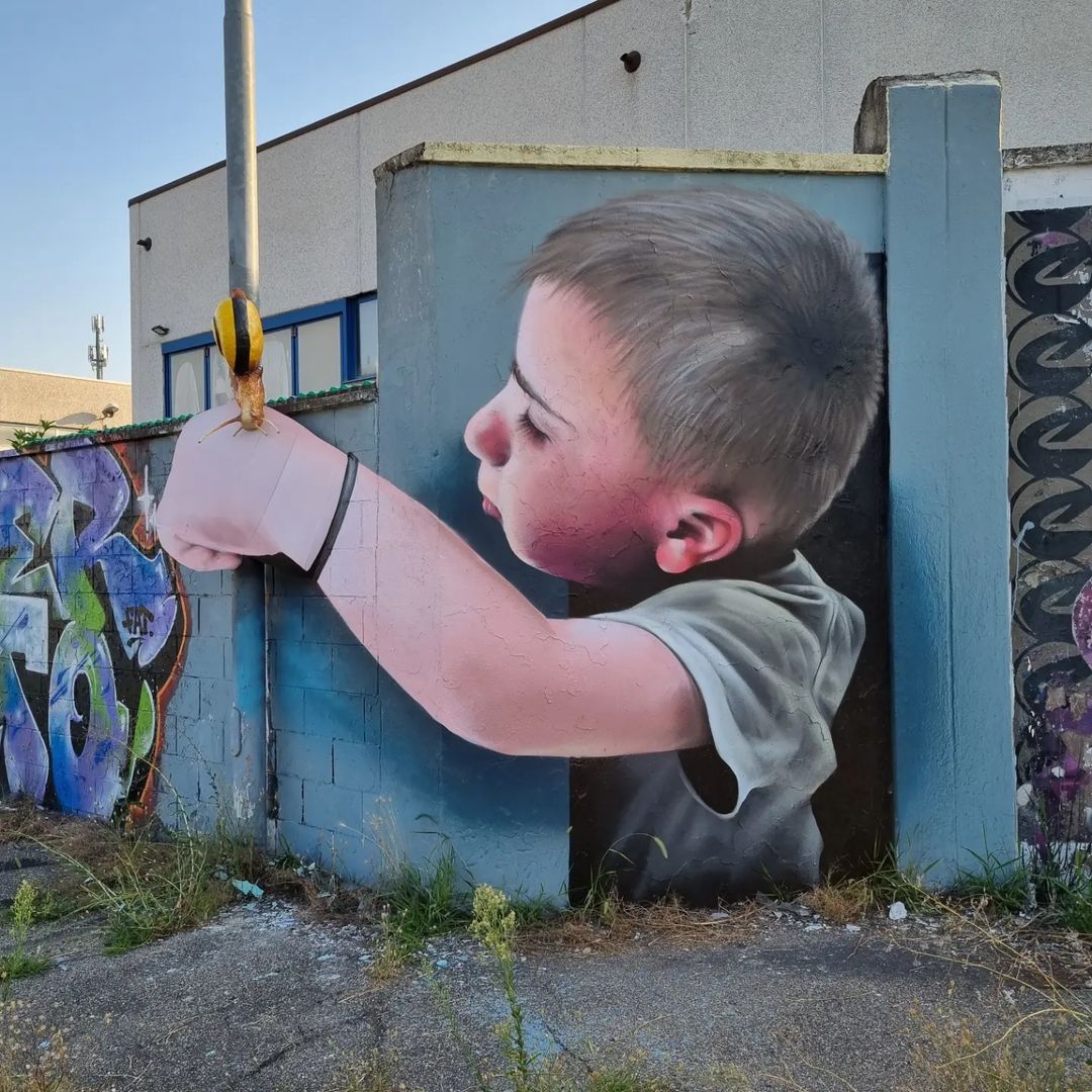 Street art mural of a boy with a focused expression reaching through a broken wall toward a yellow-black striped bollard, interacting with a real snail crawling on top, by Cheone in Italy.