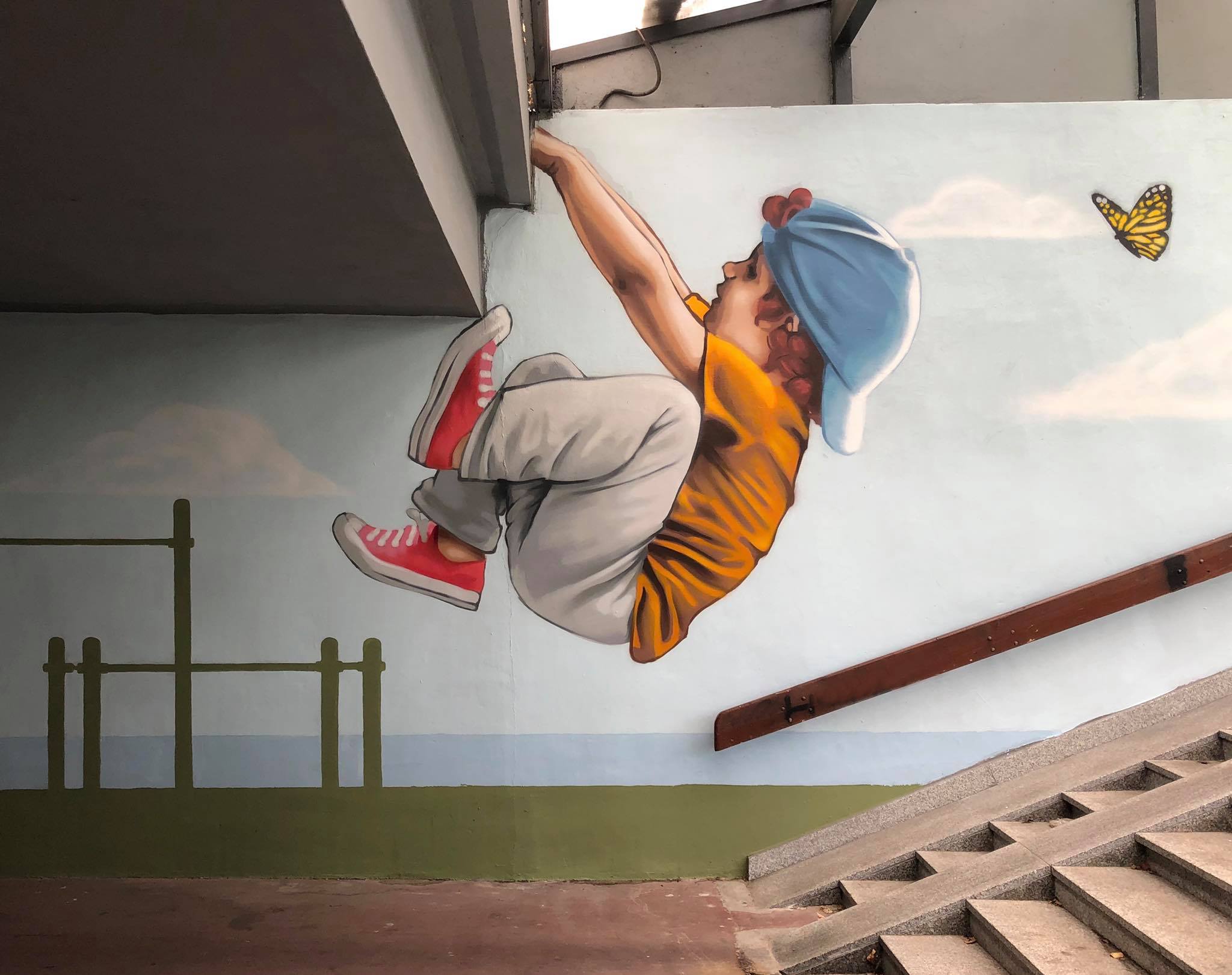 Mural of a child hanging with both hands from a building ledge, legs bent mid-swing and facing a butterfly, painted on a stairwell wall in Gdynia, Poland.