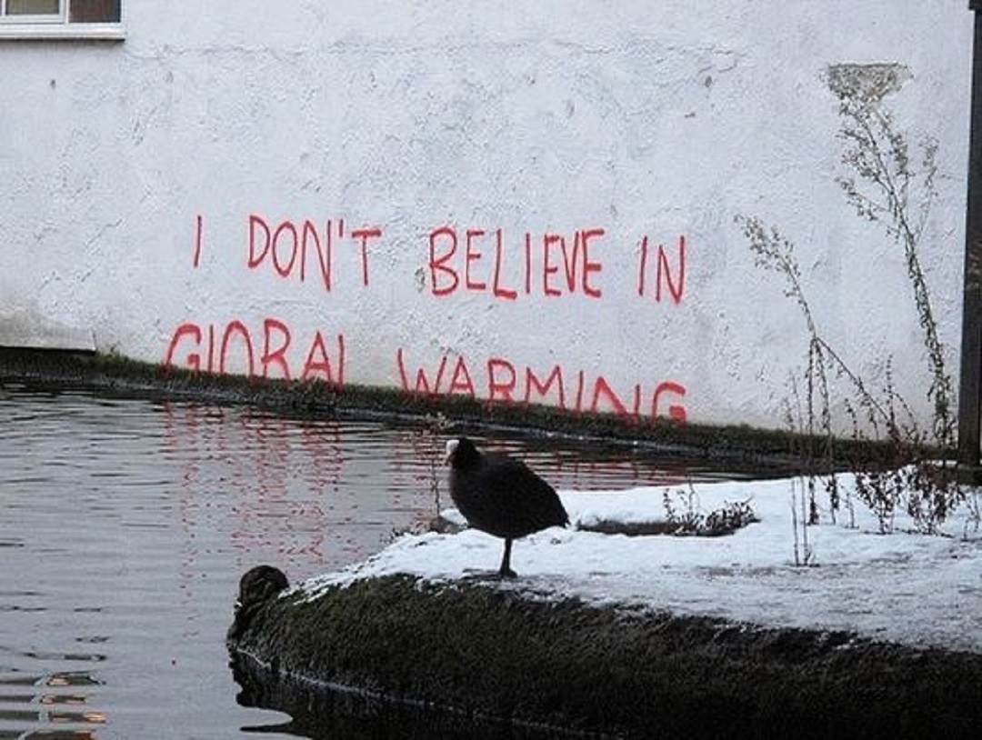Street art by Banksy showing red text on a white wall reading “I DON'T BELIEVE IN GLOBAL WARMING,” with the lower half of the words disappearing below rising canal water in London.