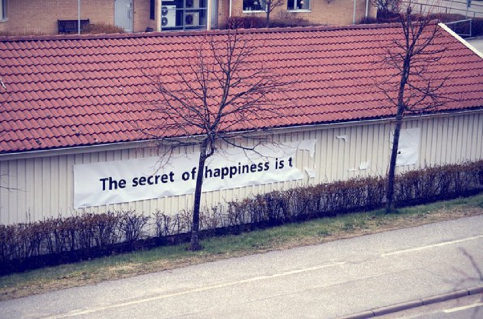Large text banner on the side of a building reads “The secret of happiness is t,” with the rest of the message torn off or missing.