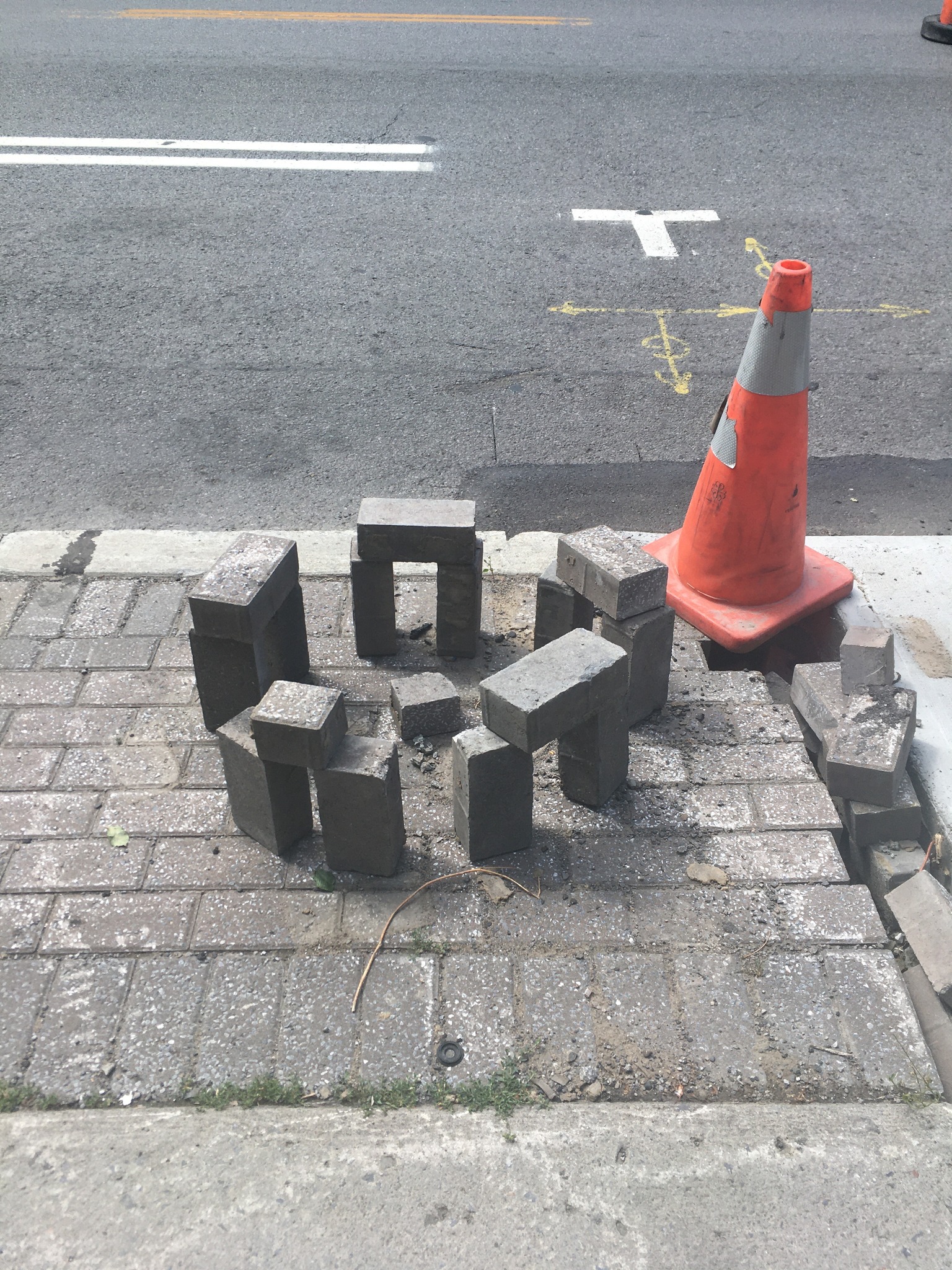 Urban paving stones stacked in a circular arrangement like Stonehenge, forming a miniature version with upright and horizontal bricks on a city sidewalk.
