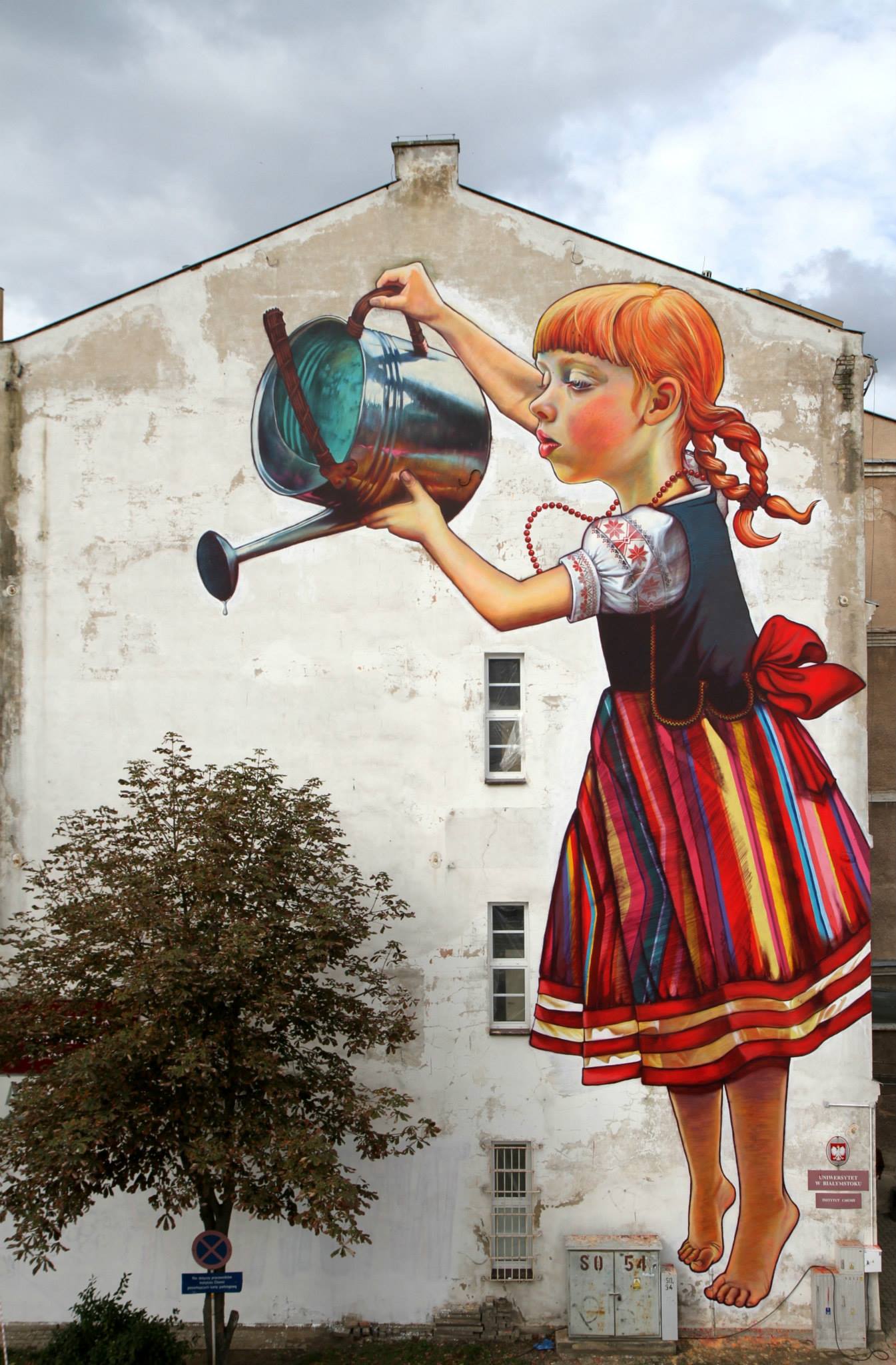 Giant mural of a girl in colorful folk clothing pouring water from a can onto a real tree that stands below the painted wall, in Białystok, Poland.