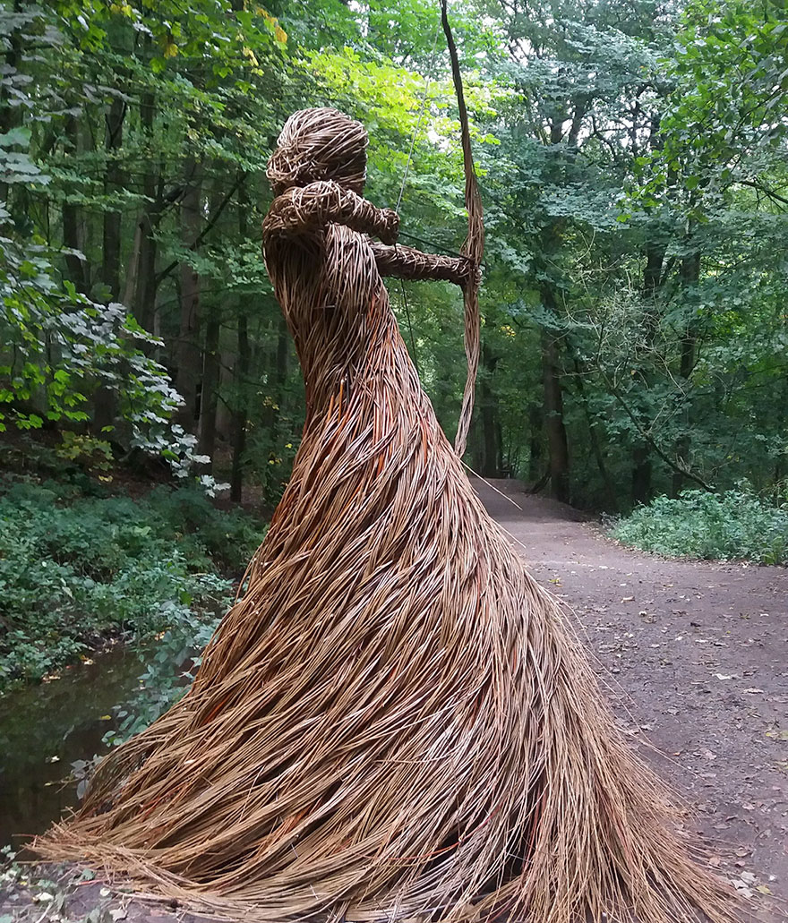 Life-sized sculpture of a woman drawing a bow, crafted from willow branches, placed in a wooded forest trail in the UK.