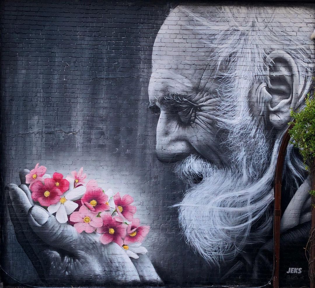 Black-and-white mural of an old man with detailed wrinkles and long white beard, holding pink flowers gently in his hands, painted by JEKS in Chattanooga, Tennessee.