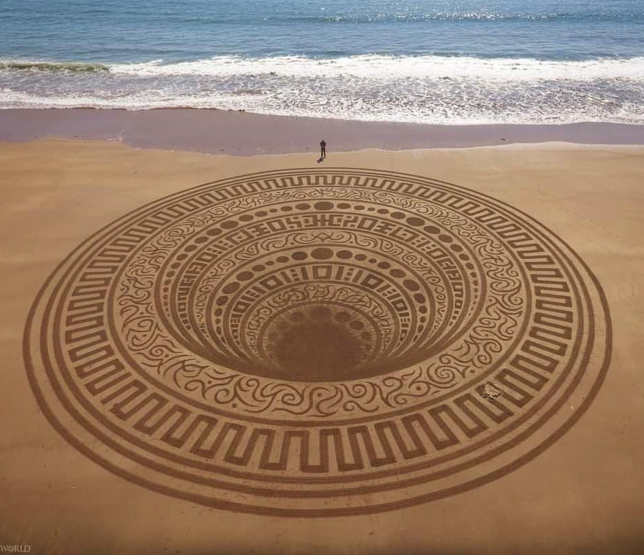 A breathtaking land art piece titled “Below” created by Jon Foreman at Lindsway Bay, Wales, UK. The design features an intricate circular pattern etched into the sand, with layers of geometric and organic motifs spiraling inward, creating the illusion of depth. A person stands near the edge, highlighting the massive scale of the artwork, with the ocean waves gently rolling in the background.
