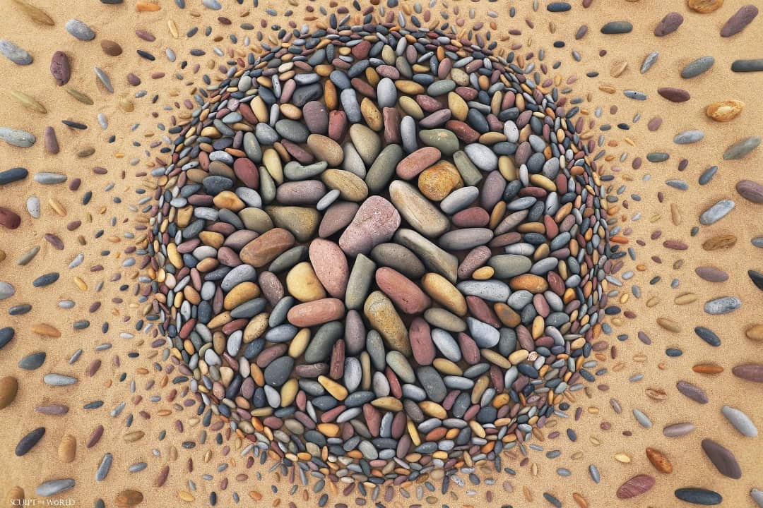 A vibrant land art piece titled “Explosia” by Jon Foreman, created at Freshwater West, Wales, UK. The design features a dense cluster of colorful stones arranged in a circular shape, radiating outward in smaller, scattered patterns. The intricate use of pebbles creates a sense of movement and energy, resembling an explosion captured in a still moment on the sandy beach.