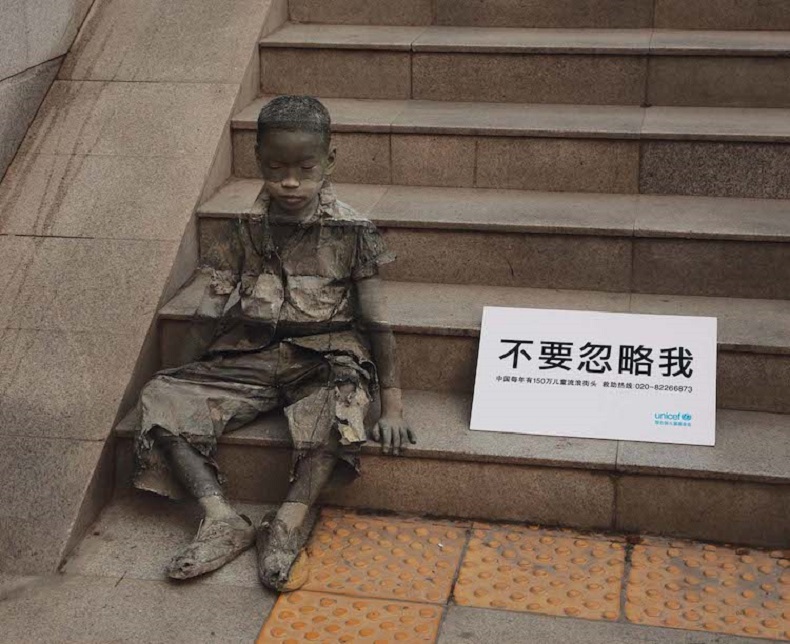 A young boy sits on a set of stone steps, painted to blend seamlessly with his surroundings as part of UNICEF China’s 2008 “Invisibility of Poverty” campaign. His tattered clothes and solemn expression evoke the silent struggles of homeless children. A white sign placed next to him displays the text “不要忽略我” (“Don’t ignore me”) in bold Chinese characters, accompanied by smaller text referencing over 1.5 million underprivileged children in China. The sign includes the UNICEF logo, emphasizing the call for awareness and action through this poignant and impactful artwork.