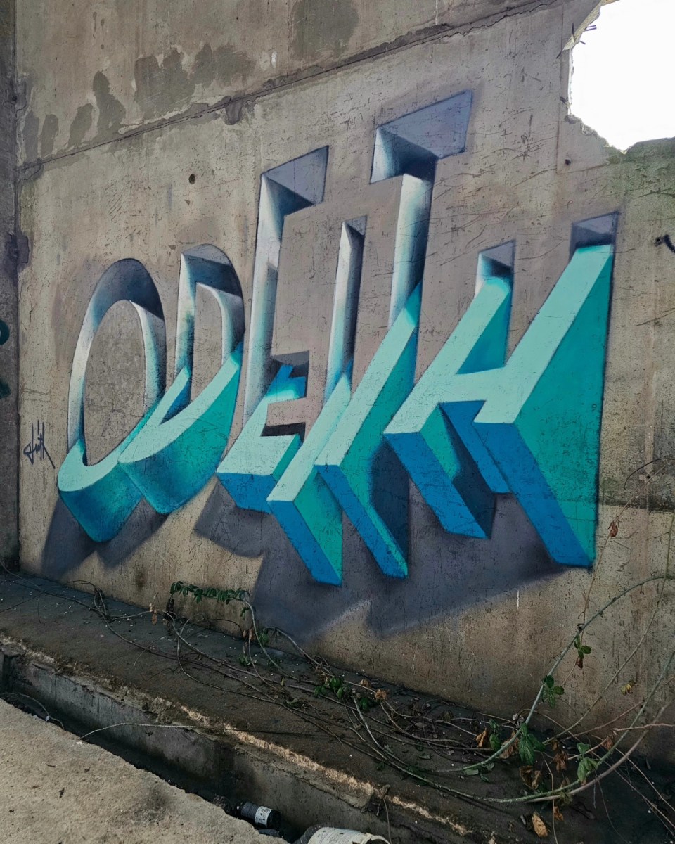 A stunning 3D graffiti piece by Odeith in turquoise tones, featuring his name written in bold, geometric lettering. The artwork appears to leap off a worn concrete wall, with expertly rendered shadows and perspective creating a mind-bending optical illusion. Surrounding the wall, overgrown vines and urban decay highlight the transformation of an abandoned space into a work of art.