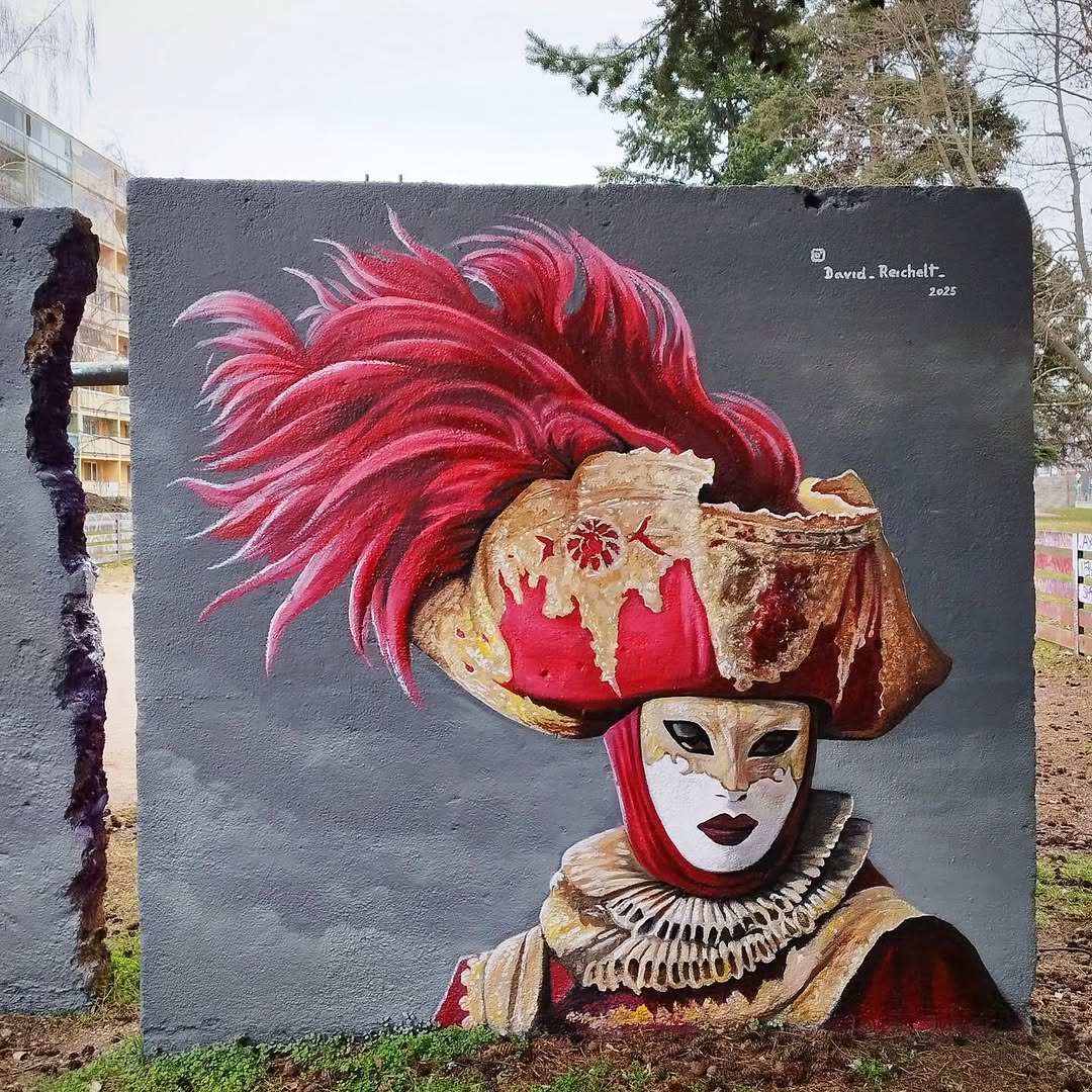 Mural of a figure in an elaborate Venetian mask and costume with a red feathered tricorn hat and golden details, painted by David Reichelt on a gray wall in Prague, Czech Republic.