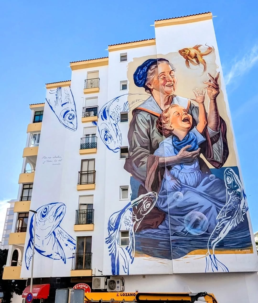 Large mural on a white building in Estepona, Spain, showing a smiling elderly woman and child reaching for a floating goldfish, surrounded by stylized blue fish, painted by Nacho Basave Cavanna.