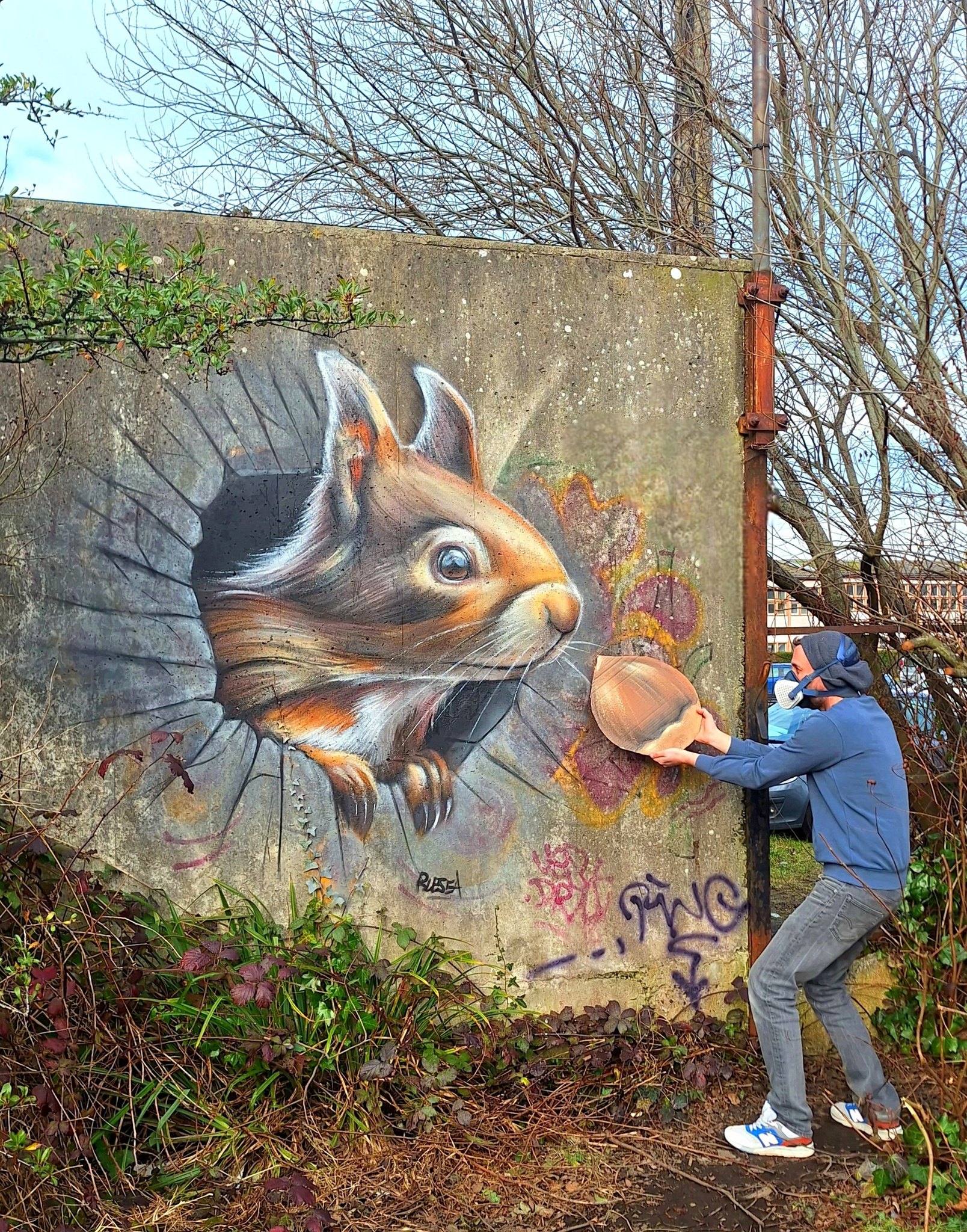 A hyperrealistic mural by Blesea in Cherbourg, France, depicting a squirrel breaking through a concrete wall. A man in a hoodie interacts with the mural, holding a real acorn, creating a playful 3D illusion.