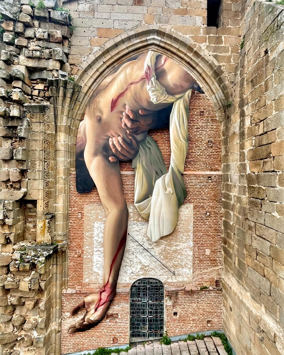 A hyperrealistic mural by J.M. Brea in Plasencia, Spain, depicting a partially draped, wounded male figure in a classical Descent from the Cross pose. The artwork is painted within the pointed Gothic arch of an old brick and stone ruin, creating a striking illusion that the figure is emerging from the building itself. Blood drips from his wounds, adding a dramatic, almost sculptural effect to the composition.