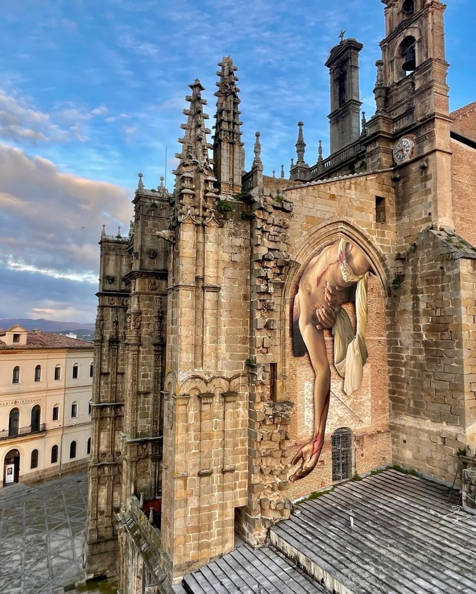 A hyperrealistic mural by J.M. Brea in Plasencia, Spain, depicting a partially draped, wounded male figure in a classical Descent from the Cross pose. The artwork is painted within the pointed Gothic arch of an old brick and stone ruin, creating a striking illusion that the figure is emerging from the building itself. Blood drips from his wounds, adding a dramatic, almost sculptural effect to the composition.