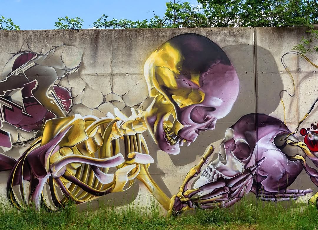 A mural by SEAROK in Freital, Germany, depicting a large, semi-abstract skeleton painted in shades of gold, purple, and black. The skeletal figure appears to be emerging from the cracked concrete wall, holding another skull in its hands in a dramatic and almost theatrical pose. The highly detailed rendering of the bones contrasts with graffiti-style lettering on the left side, adding an urban, surreal touch to the piece.
