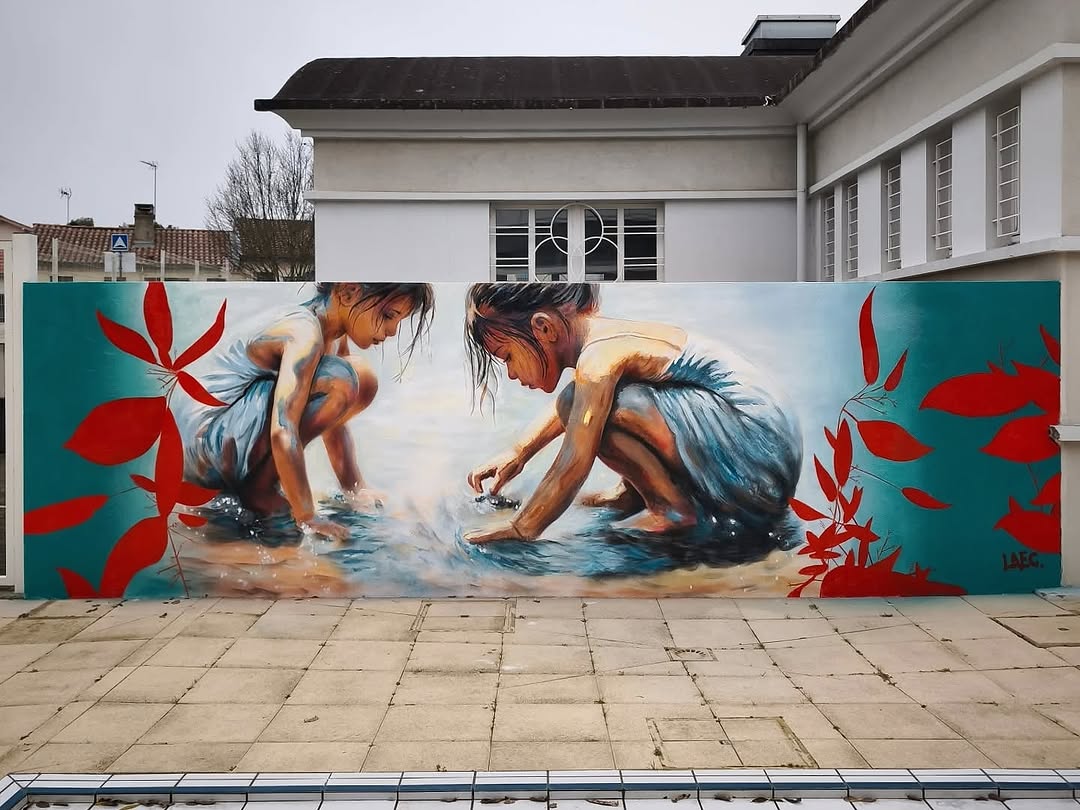 A mural by LAEC in Mont-de-Marsan, France, depicting two young girls in blue dresses crouching by a shallow pool of water, playing and exploring. The hyperrealistic artwork is accented with bold red leaves against a teal background, adding a dreamlike, painterly contrast. Their wet clothes and glowing skin reflect the sunlight, making the scene feel alive and full of nostalgia.