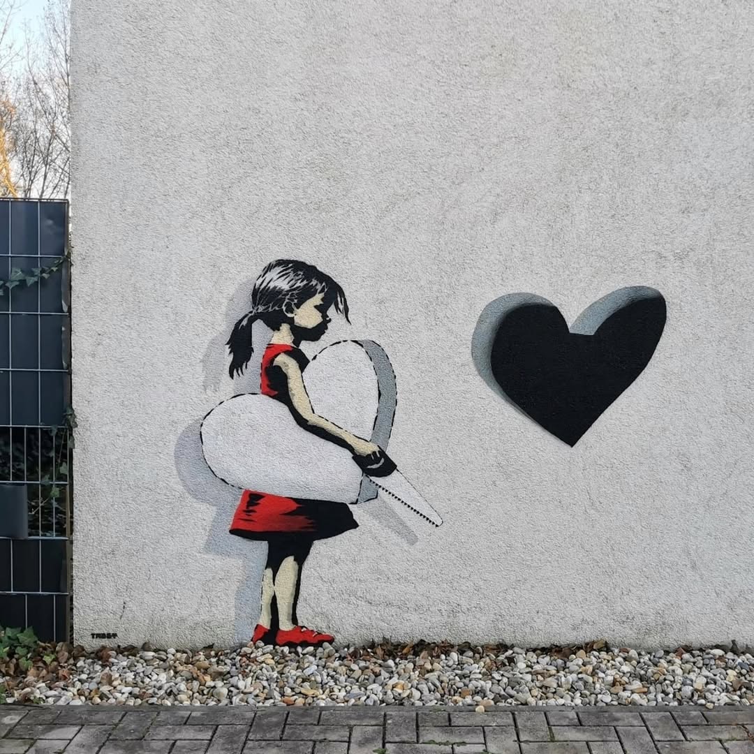 A stencil street art piece by TABBY featuring a young girl in a red dress holding a white heart with a chainsaw in her hands. To her right, a separate black heart appears cut out from the wall, casting a shadow that adds a 3D effect. The artwork plays with perspective and symbolism, evoking themes of love, loss, and creation.