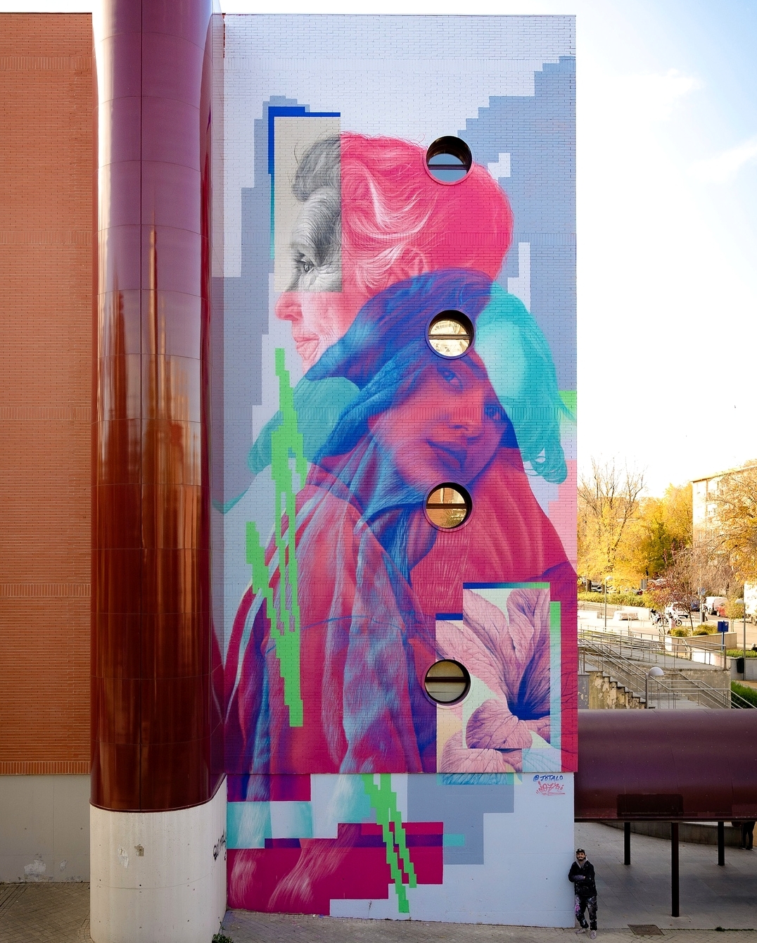 A large-scale mural by Jota López in Madrid, Spain, titled El Abrazo. The artwork features two overlapping portraits—an elderly woman in grayscale and a younger woman in vibrant shades of pink, blue, and purple. Digital pixelation and glitch-like effects fragment the composition, blending tradition with modernity. Circular windows on the building integrate seamlessly into the design, enhancing the illusion. A small figure stands beneath the towering mural, emphasizing its scale.