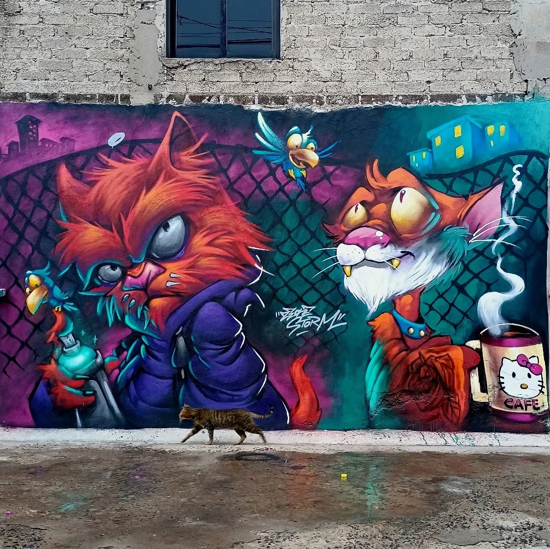 A colorful mural by Zeon and Luis Zúñiga in Mexico City, depicting two anthropomorphic cats with exaggerated expressions. On the left, a tough, streetwise cat in a hoodie grips a spray can, with narrowed eyes and sharp fur. On the right, an older, disheveled cat with a white beard and wide eyes sips from a steaming coffee mug adorned with a Hello Kitty design. The background features a chain-link fence, flying cartoonish birds, and neon city lights, adding to the comic-book-style energy of the piece. A real cat casually strolls past, blending into the scene.