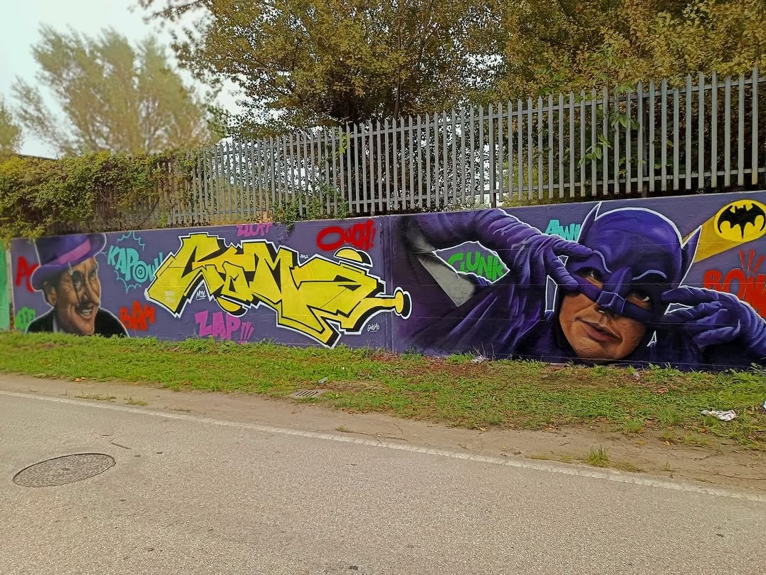 A colorful mural by Matteo “Ilcoffee” Fronduti in Bastia, Italy, featuring a playful rendition of Batman and The Penguin, surrounded by graffiti-style lettering and classic comic book sound effects like 'Kapow!' and 'Zap!!!'. Batman, in a deep purple suit, makes a hand gesture as if adjusting his mask, while The Penguin smirks mischievously in his signature hat and monocle. The bright yellow Batman logo pops against the dynamic, nostalgic design, blending street art with pop culture.