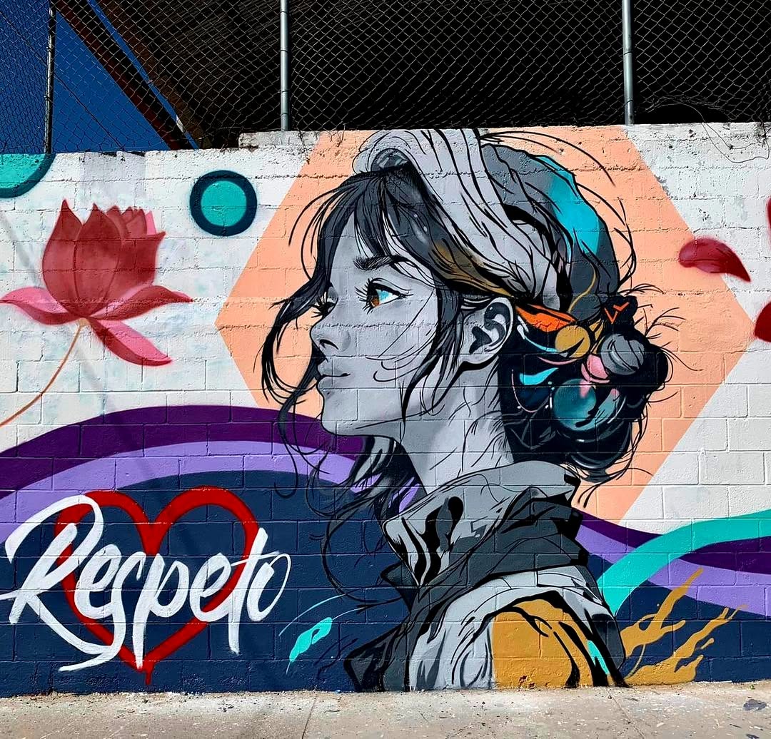 A mural by Mick in Ciudad Juárez, Mexico, featuring a grayscale portrait of a young woman with colorful highlights in her hair, wrapped in a headscarf. Her gaze is strong and serene, set against a geometric pastel backdrop with a blooming lotus flower and a heart-shaped ‘Respeto’ (Respect) message in elegant script. Flowing purple and blue waves add movement, symbolizing harmony and empowerment.