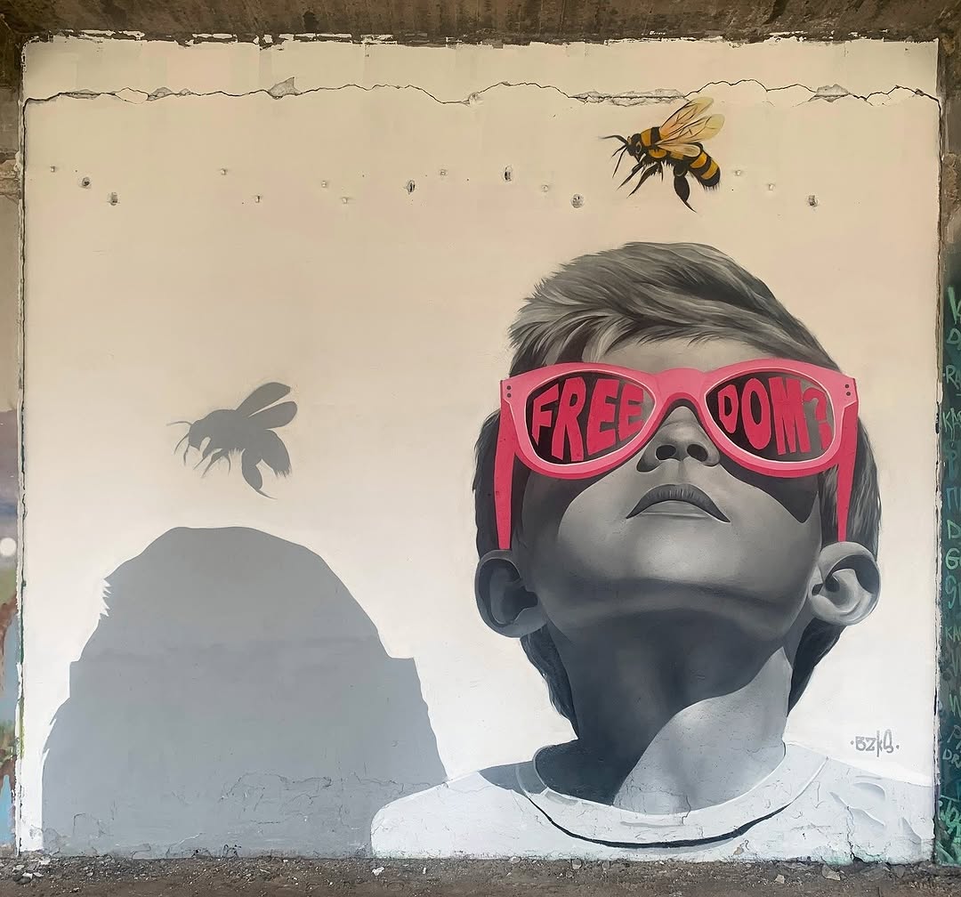 A mural by BZKS in Thessaloniki, Greece, depicting a grayscale portrait of a young boy wearing oversized pink sunglasses with the word 'FREEDOM?' written across the lenses. The boy gazes upward at a realistic bee hovering above, while a second bee appears only as a shadow on the wall. The contrast between the monochrome face and the vibrant sunglasses adds depth and symbolism, inviting reflection on freedom and perspective.