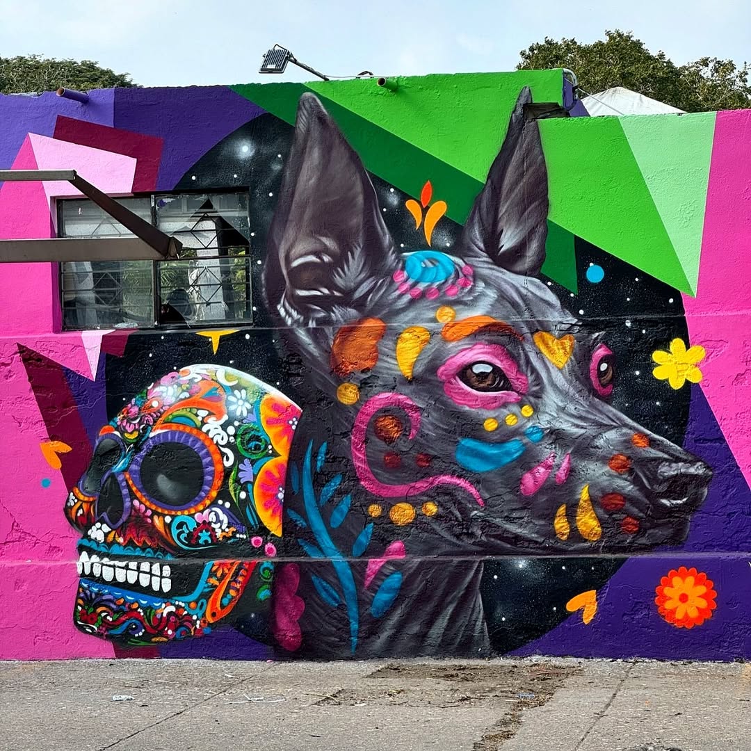 A mural by Juan Pablo and Adán Yadir Rodríguez in Tampico, Mexico, featuring a Xoloitzcuintli dog painted in a hyperrealistic style with colorful Día de los Muertos-inspired decorations on its face. Beside it, a sugar skull adorned with intricate floral patterns and neon hues glows against a cosmic black background. The mural is framed by geometric shapes in bright pink, green, and purple, adding a modern, abstract touch to the traditional Mexican symbolism.