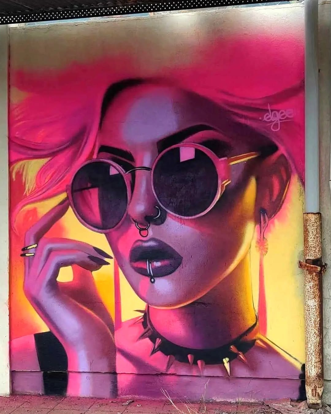 A striking mural by Elgee featuring a neon-lit portrait of a woman with bold piercings, dark lipstick, and oversized round sunglasses reflecting the surrounding world. Her hair flows in shades of hot pink and yellow, blending seamlessly into the background. A spiked choker and sharp nails add an edgy, rebellious touch, making the piece radiate with attitude and intensity.