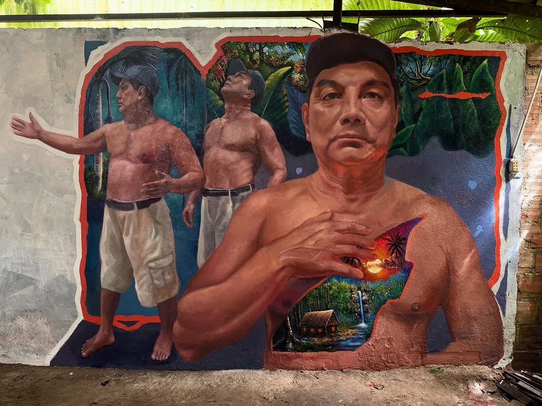 A mural by Diego Capuena in Yurimaguas, Peru, featuring three portraits of Manuel Capuena set against a lush jungle backdrop. The largest figure in the foreground places a hand over his chest, where a vivid scene of a tropical landscape, complete with a thatched-roof hut, waterfall, and sunset, is painted within his body. The artwork captures themes of identity, ancestry, and the deep connection between humanity and nature.