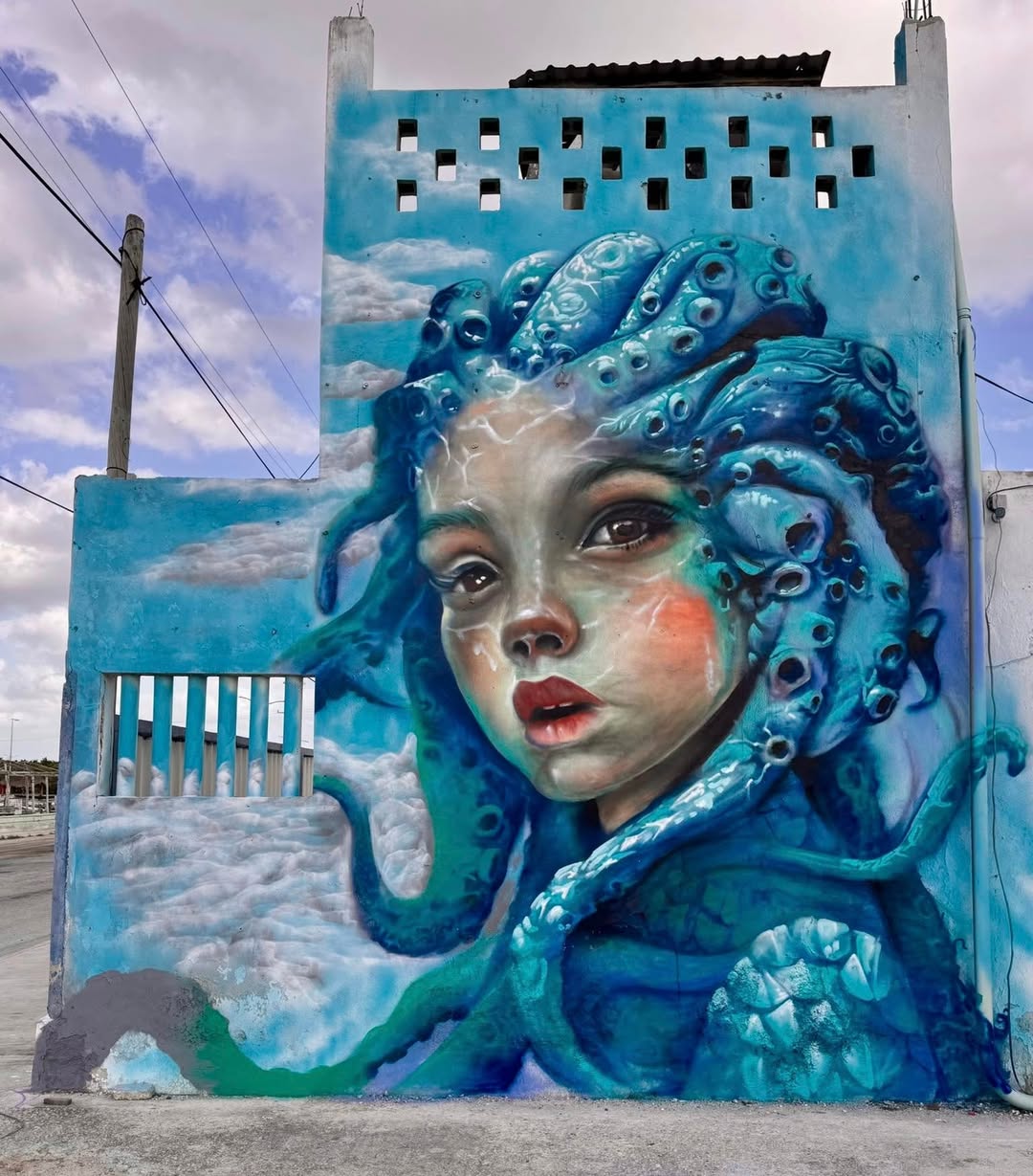 Mural depicting a girl's face with octopus tentacles as hair, painted in shades of blue and green, set against a cloudy sky backdrop in Río Lagartos, Mexico.