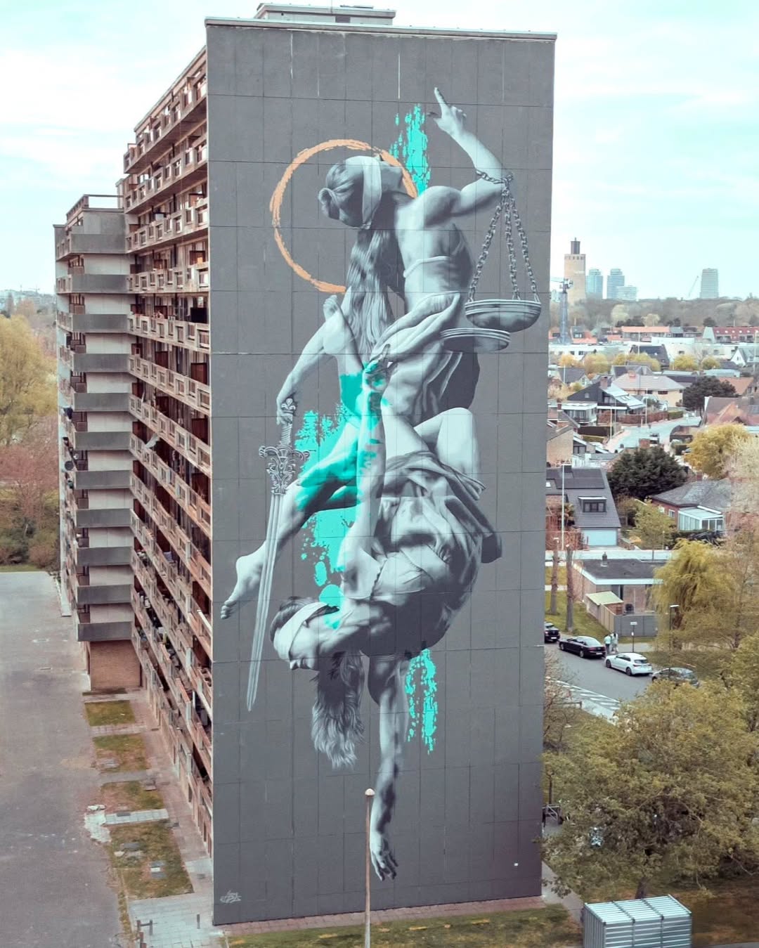 Large-scale mural of an upside-down Lady Justice holding scales and a sword, detailed in grayscale with turquoise and gold highlights, painted on a tall building in Ostend, Belgium.