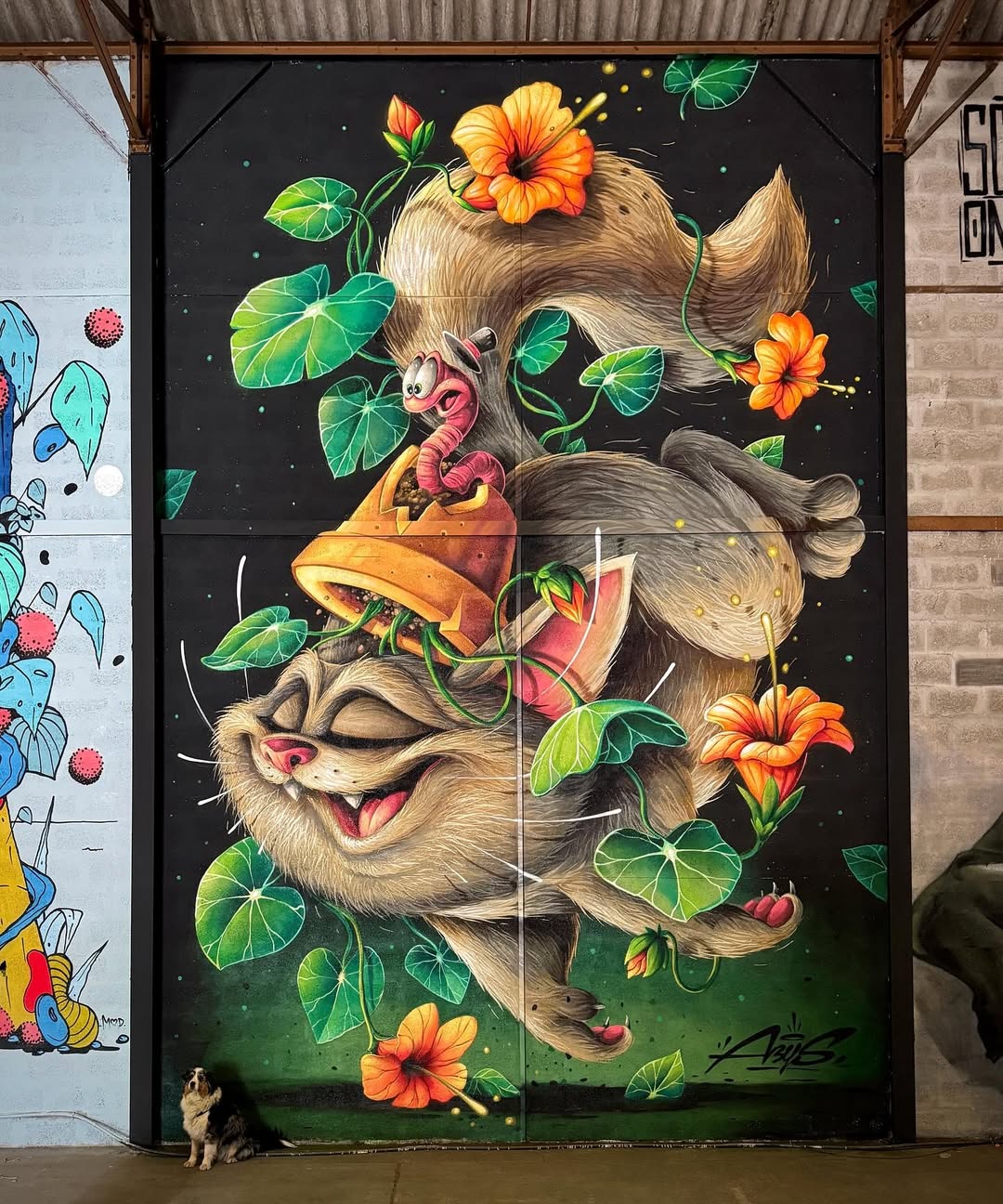 Whimsical mural featuring a smiling cat tumbling through bright flowers and greenery, accompanied by a cartoonish worm character in a flowerpot hat, painted on a black background in Bourgoin-Jallieu, France.