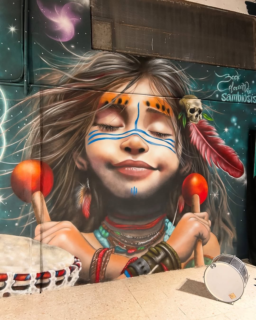 Mural of a smiling indigenous girl holding maracas, with traditional face paint, feather adornments, and a vibrant atmosphere of cultural celebration, painted in Granada, Spain.