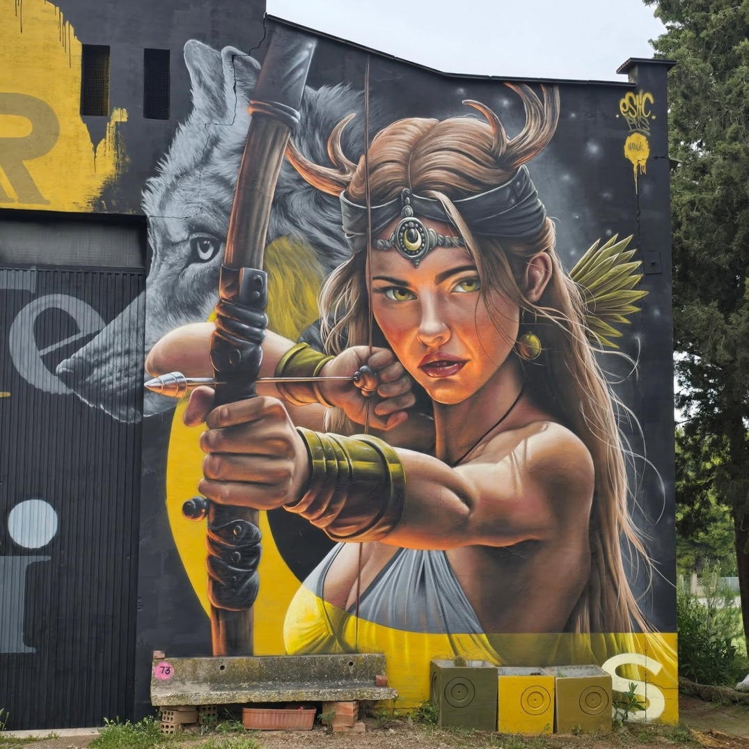 Mural of a fierce female archer aiming her bow, with detailed clothing and jewelry, and a wolf in the background, painted on a large building wall in Penelles, Spain.