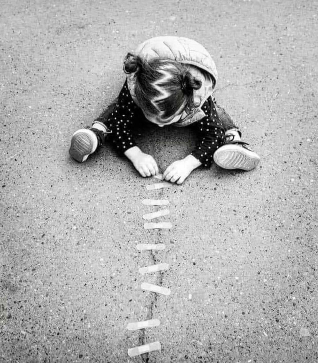 Street art photograph of a child wearing a puffy vest sitting cross-legged on cracked asphalt and placing oversized cartoon bandages across the crack.