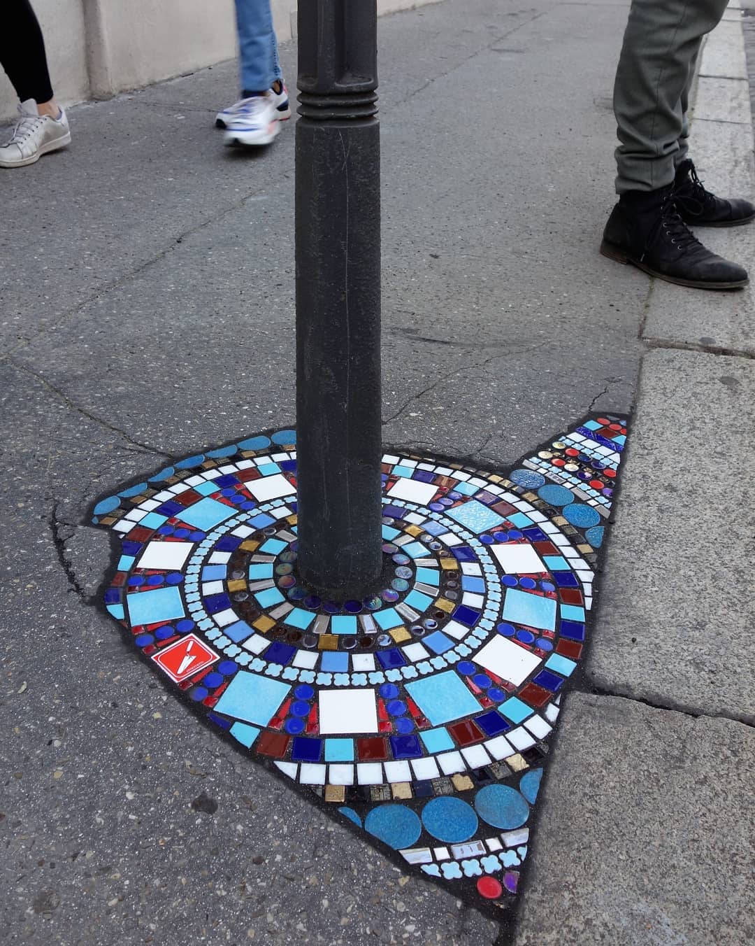 Broken asphalt around a lamppost filled with vibrant mosaic tiles by Ememem in Lyon, France, arranged in a radial pattern with geometric shapes and deep blue accents.