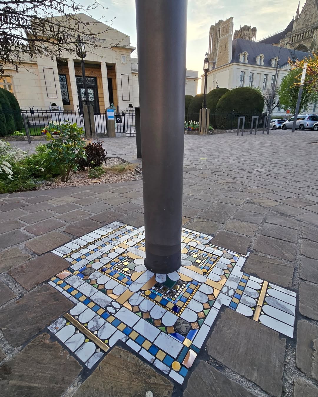 Mosaic artwork surrounding a streetlamp pole in Reims with white petal shapes and golden tiles embedded into the cobblestone.